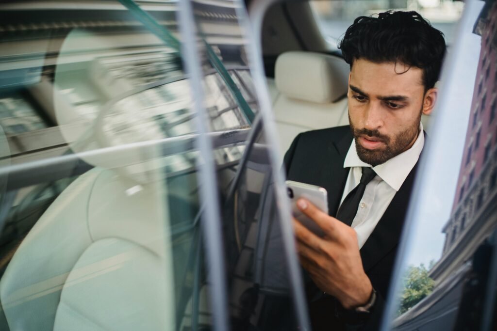 Confident businessman in suit using smartphone inside car, viewed through window reflection.
